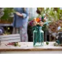 Bunter LEGO-Blumenstrauß in transparenter grüner Vase auf Holztisch, mit unscharfer Person im Hintergrund Bunter LEGO-Blumenstrauß in transparenter grüner Vase auf Holztisch, mit unscharfer Person im Hintergrund