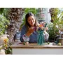 Une femme souriante arrange un bouquet de fleurs LEGO colorées dans un vase sur une table en bois dans un jardin verdoyant