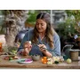 Une femme assemble des fleurs LEGO colorées à une table en bois avec des pièces éparses et un arrosoir vert