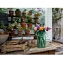 Bouquet LEGO coloré dans un vase vert transparent posé sur une table en bois dans une serre avec des plantes en pots