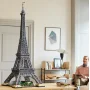 Maquette LEGO de la tour Eiffel gris foncé avec base verte, exposée dans un salon moderne à côté d'un homme assis